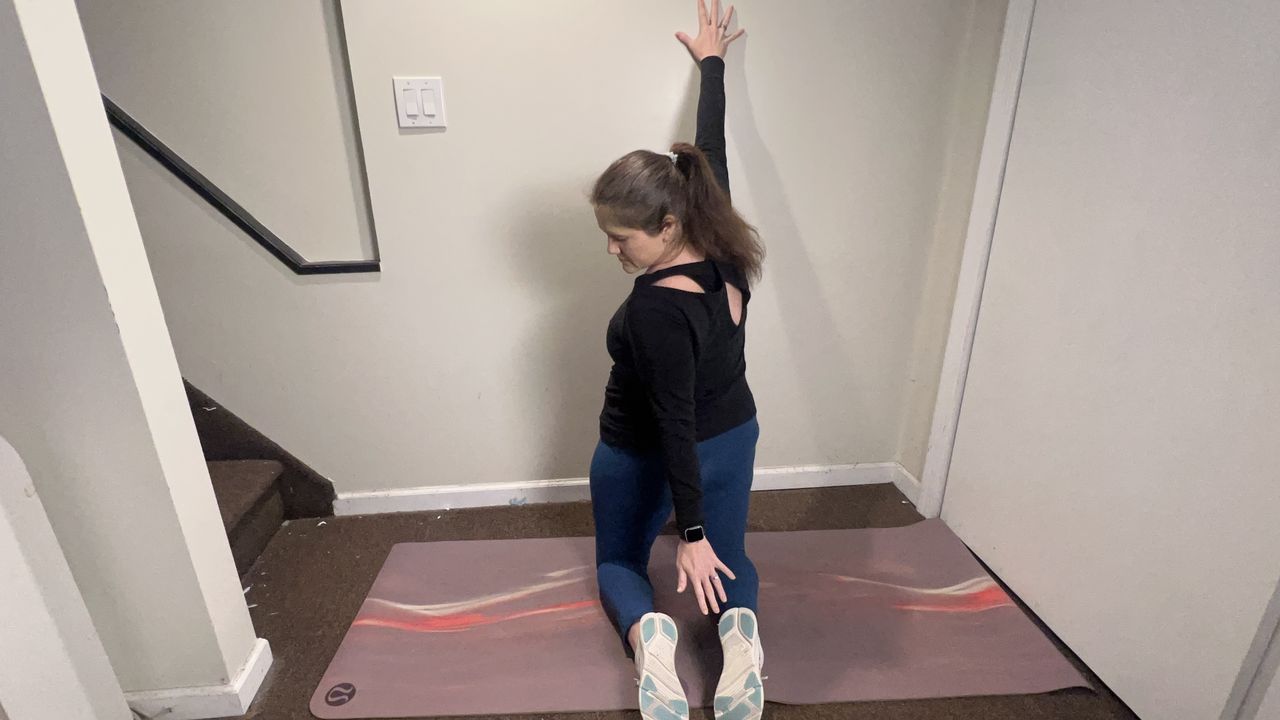 Trainer Jennifer RIzzuto performs a back mobility stretch on the floor. She is kneeling close to a wall on a yoga mat, right hand on the wall and left hand reaching back towards her right heel.