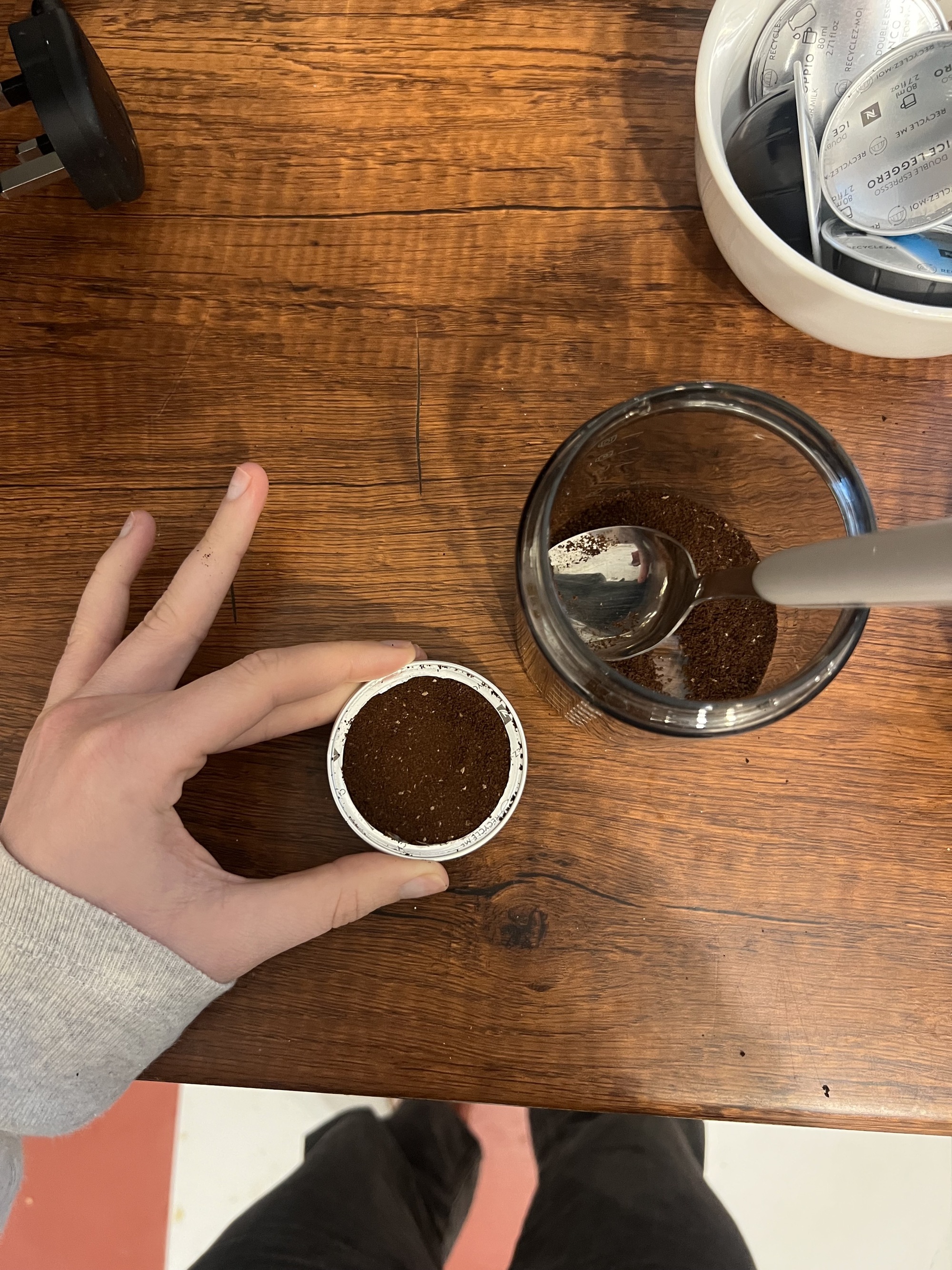 image of a hand wearing a long-sleeved gray shirt holding an open coffee pod filled with fresh grounds over a wooden countertop. There is a jar of coffee ground with a spoon in it and a bowl of coffee pods also on the counter.