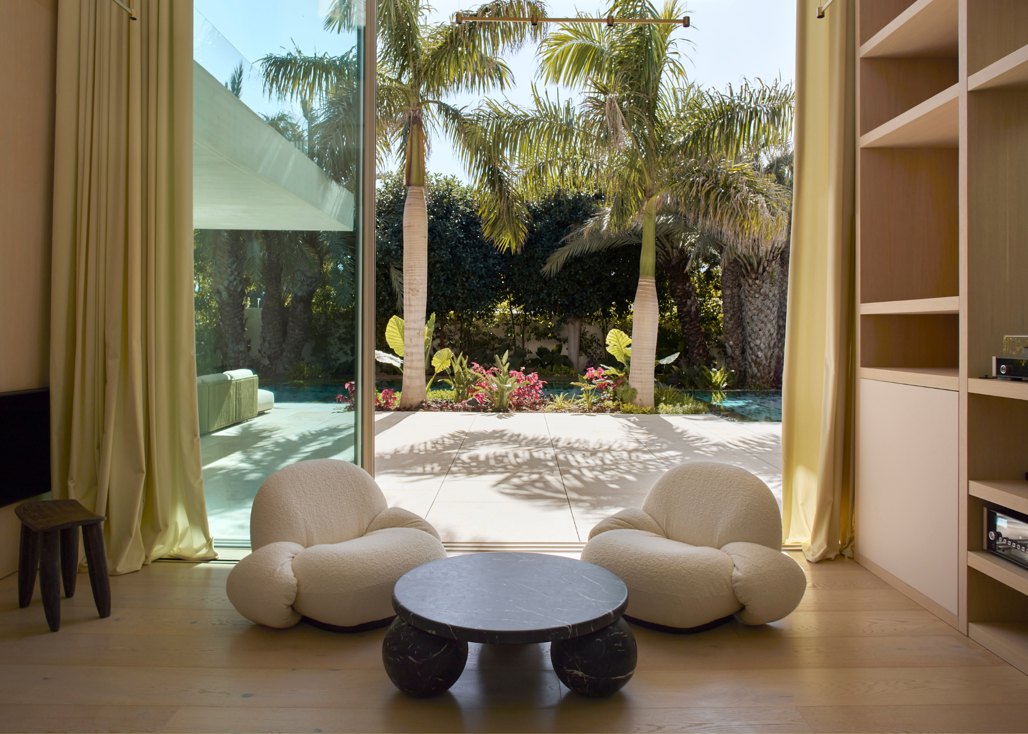 A living room with doors to a tropical backyard. Indoors are a pair of white armchairs, a marble coffee table with bobbin feet, and a wood stool
