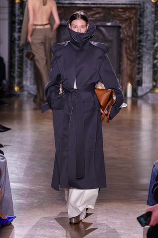 a model walks the victoria beckham runway wearing a trench coat with an ultra high neck