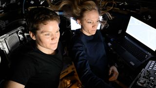 two women in dark clothing float beside each other inside a windowed module of a space station