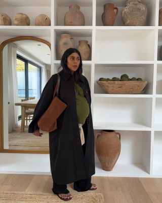 Woman wearing Sezane sweater, black trench coat, black pants, flip-flops, and brown suede bag.