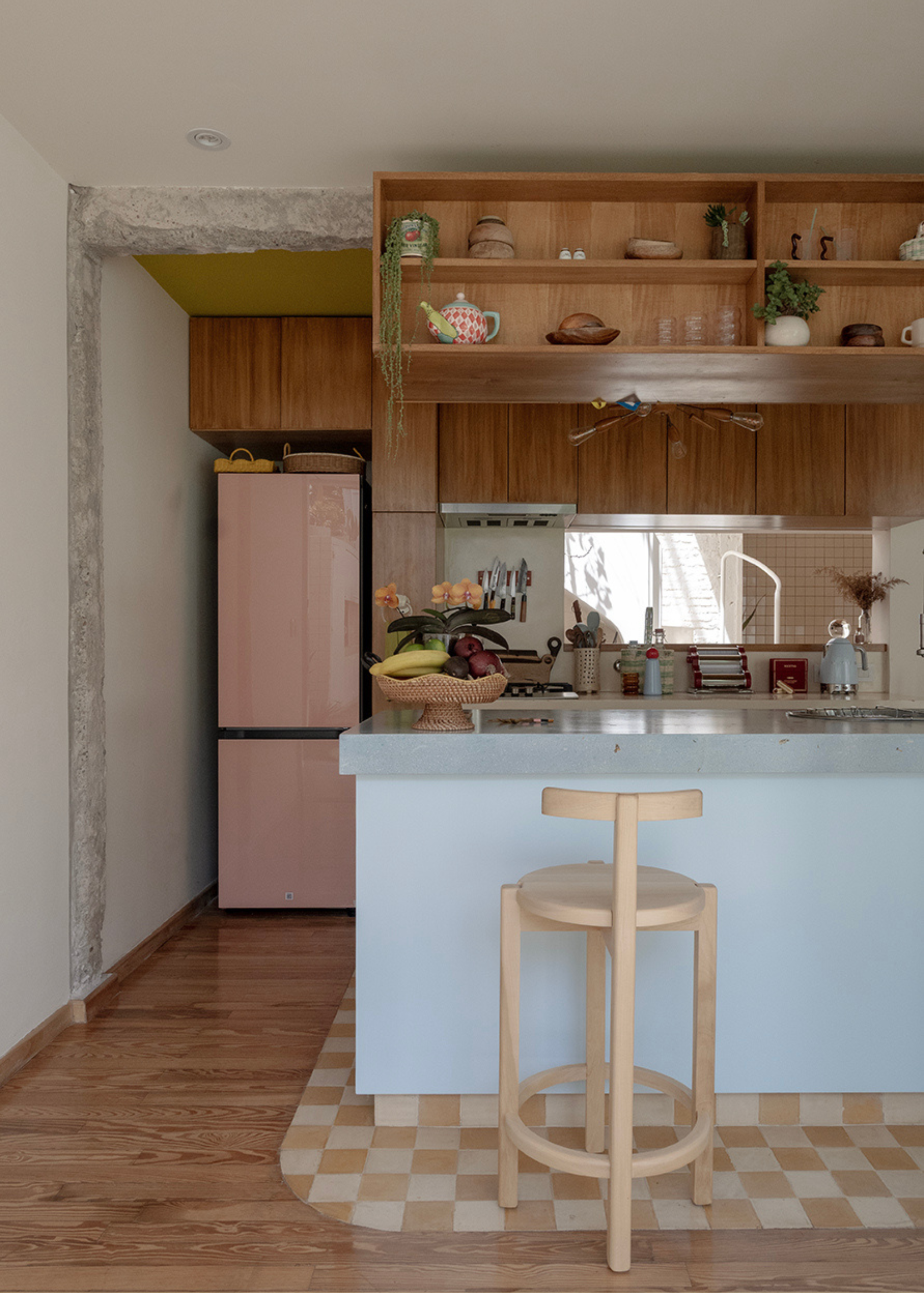 A kitchen with wooden cabinets, a floating wooden shelf, a pale blue kitchen island with a neutral border tile finish and a wood stool