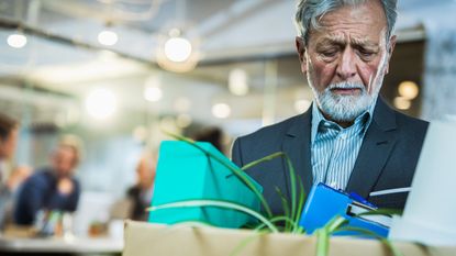 An older man carries a box of belongings after losing his job near retirement.
