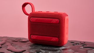 The back face of a red JBL Go 4 pictured against a pink background. It is sitting on a dark stone surface with puddles of water around it.