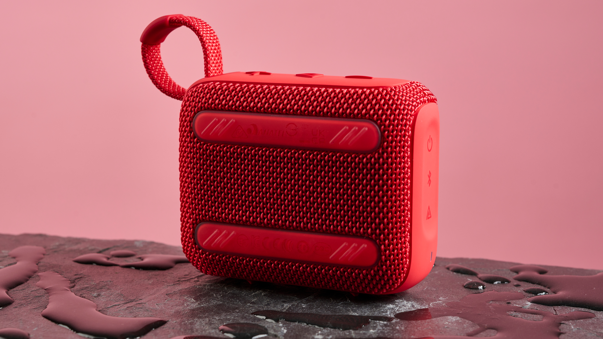 The back face of a red JBL Go 4 pictured against a pink background. It is sitting on a dark stone surface with puddles of water around it.