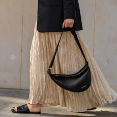 a close up photo of a woman wearing minimal sandals with a tan pleated dress and black crescent bag