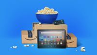 An Amazon Fire tablet and Fire TV Stick arranged with Amazon boxes and a bowl of popcorn on a blue background. 