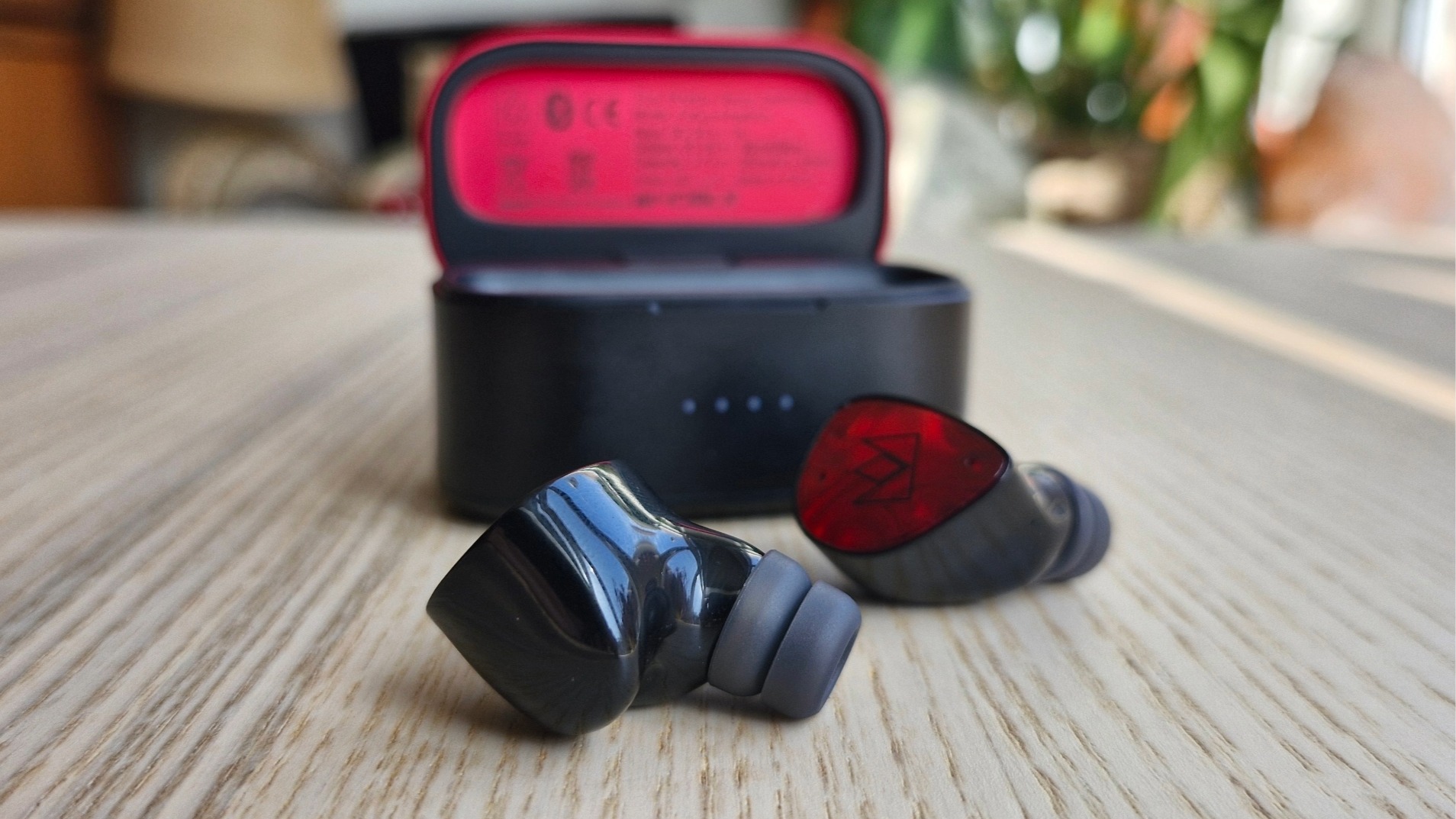 Noble FoKus Amadeus wireless earbuds, in front of their carry case, on a wooden surface