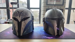 The Black Series Bo-Katan Helmet (left) and the Denuo Novo The Mandalorian Helmet (right) side-by-side, facing forwards. The Bo-Katan Helmet has a gray, black, and white owl-like pattern on the light blue helmet and a black T-shaped visor. The Mandalorian Helmet is just slightly smaller in size, a shiny metallic, gray and has a more straight T-Shaped black visor.