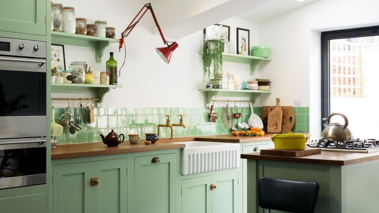 Green kitchen with bright window on right and butler sink, with open shelving and double stacked oven