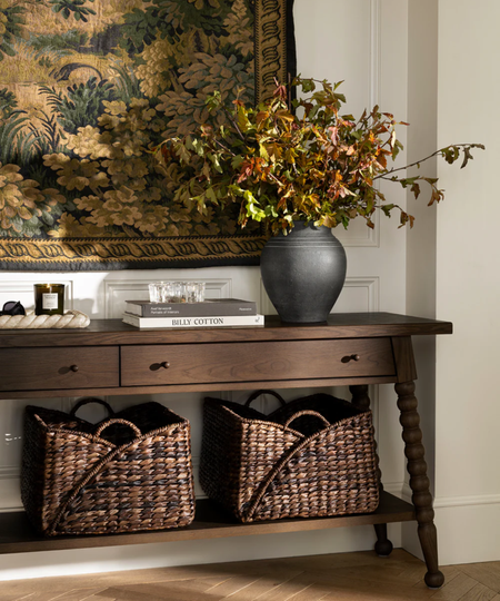 a credenza with two woven storage baskets on the bottom shelf, a vase of flowers on the top shelf, and a tapestry hanging from the wall