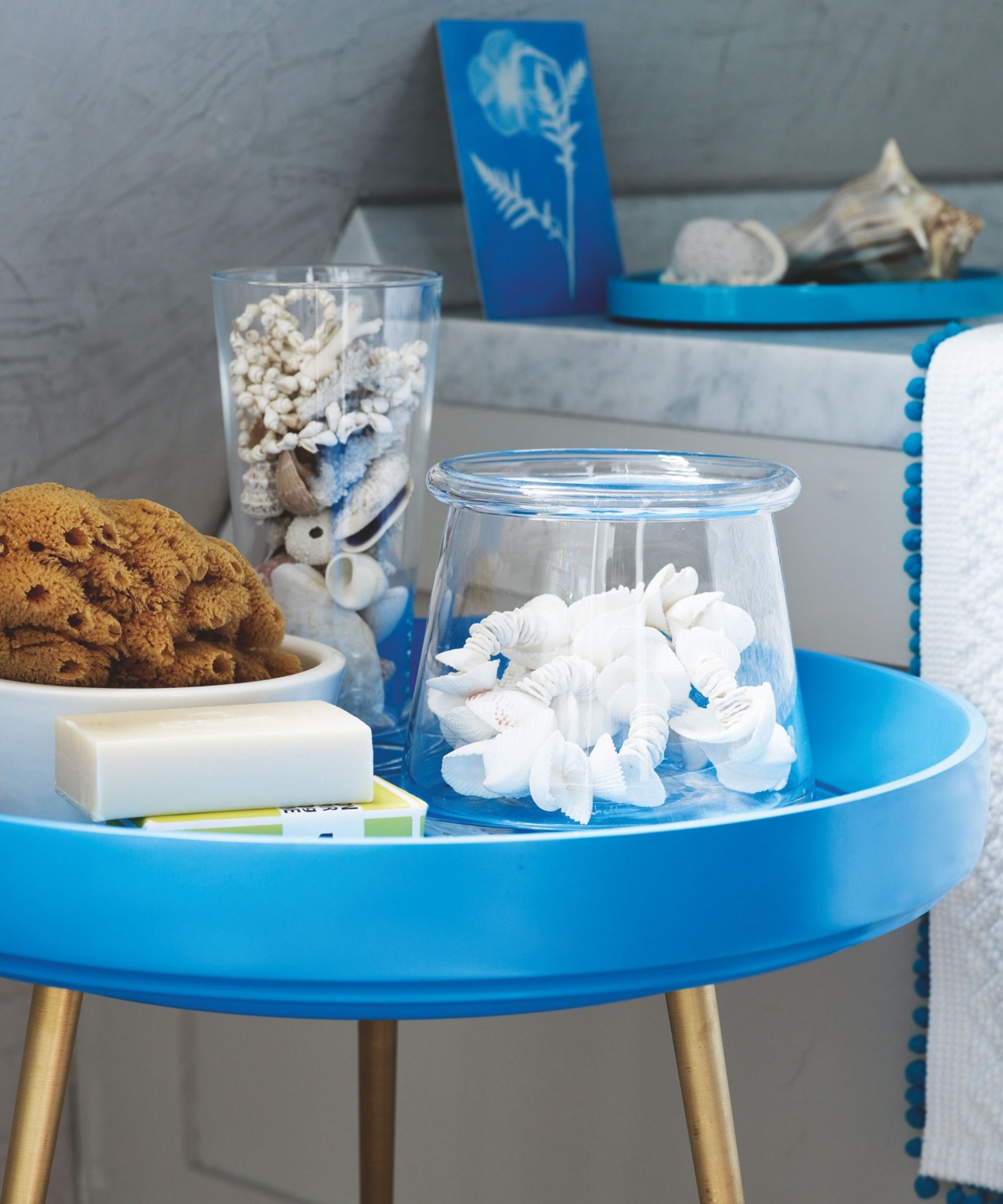 Nautical bathroom blue table with gold legs and shells in glass vases. Behind is a marble countertop with shells and cyanotype.