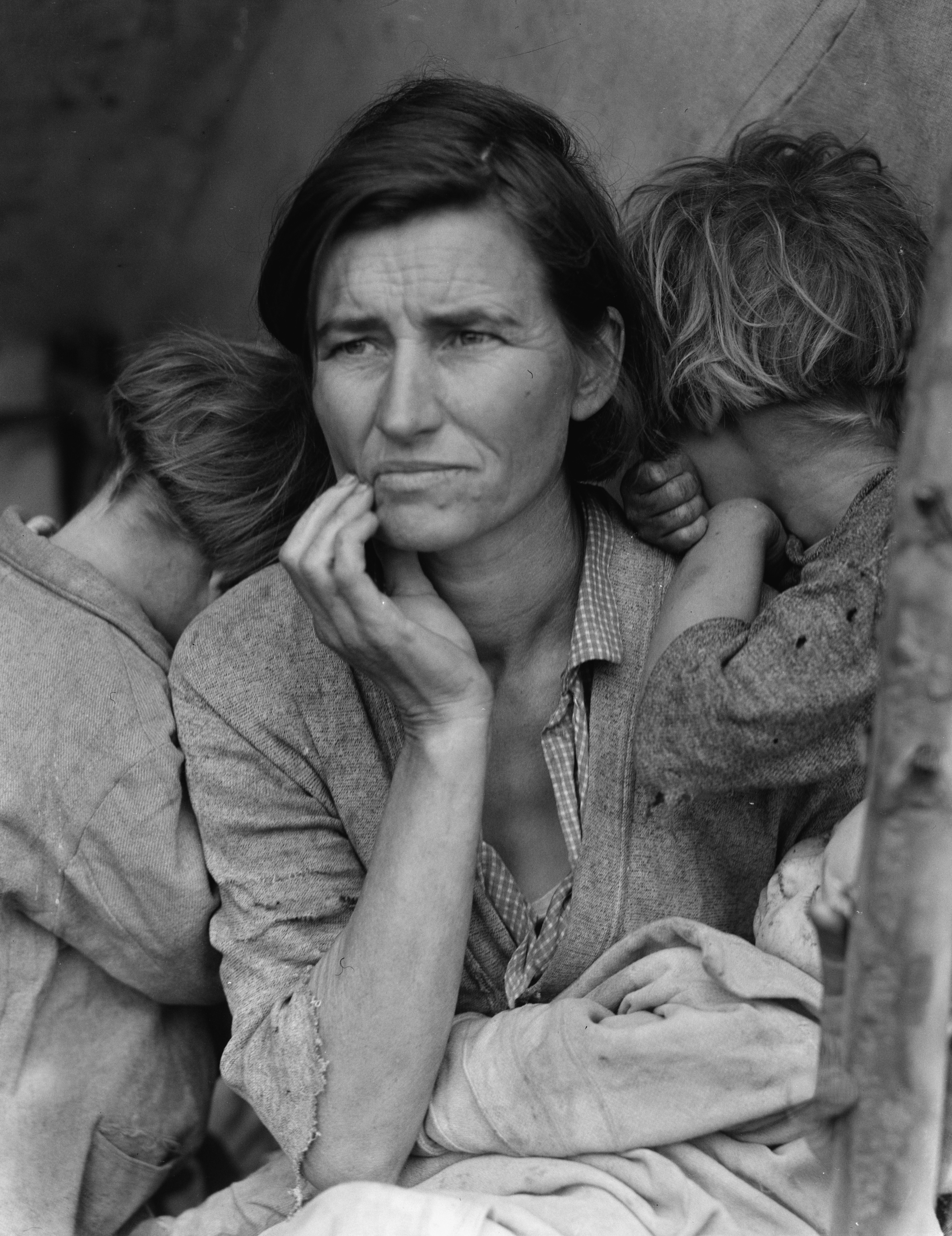 A careworn mother rests her chin on her hand, gazing into the distance while two children bury their faces against her shoulders.