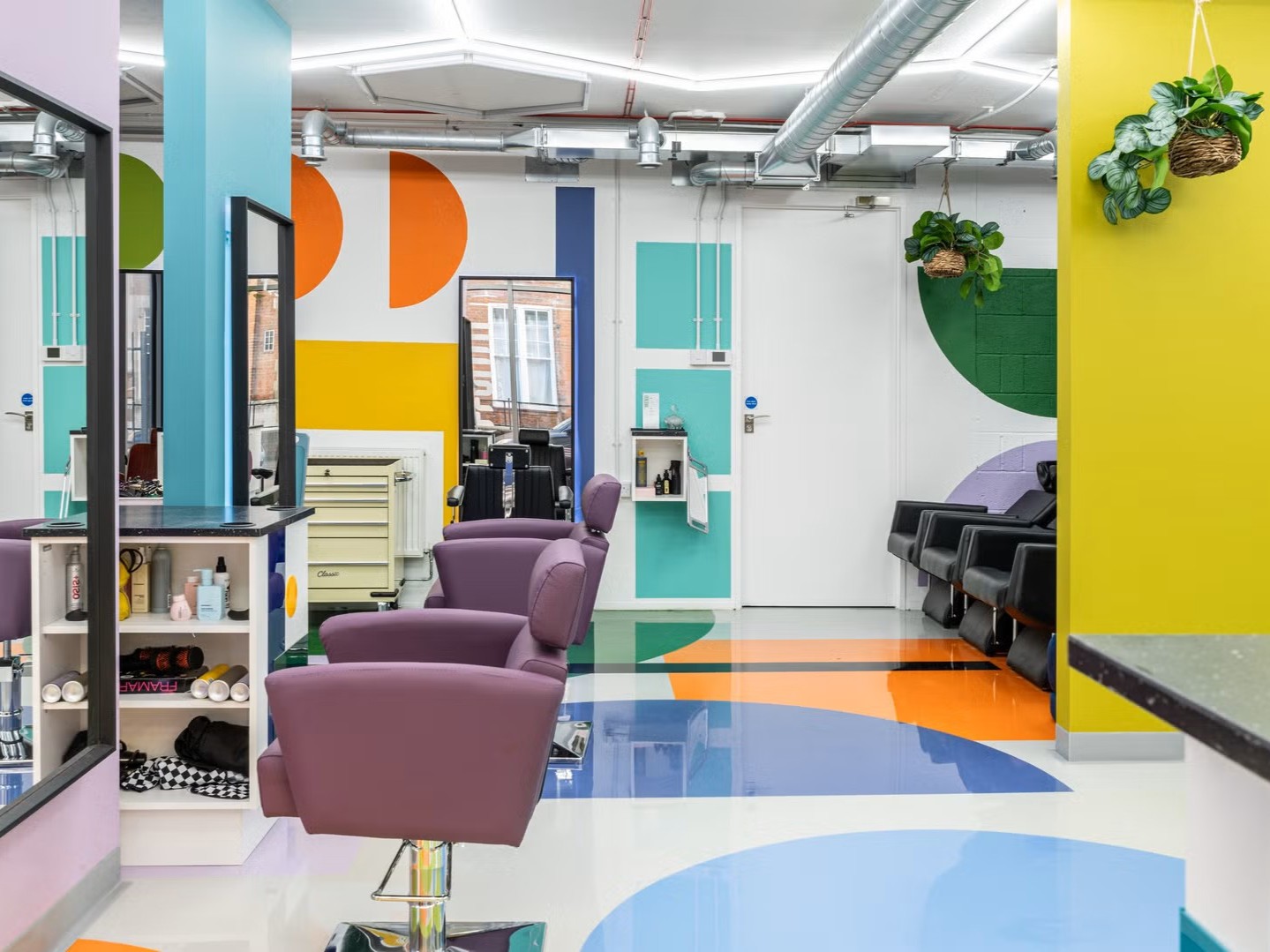A colorful, neon-tinted hair salon with geometrical shapes on floors and walls, and quirky, sculptural furniture in purple, red, yellow, and baby blue.