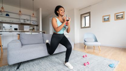 A woman performs a Bulgarian split squat on a couch at home. She is positioned in front of the couch, with one foot resting behind her on the couch and the other on the floor. Her knee is bent as she moves down into the squat and she holds a dumbbell in her hands at chest height. We see a kitchen behind her, shelving, decorative lights and a chair.