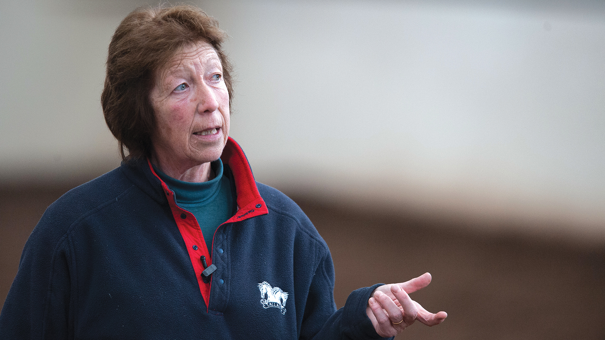 Dressage rider and trainer Pammy Hutton wears a navy top with a red collar, is looking out of the image to her left and is making an open hand gesture