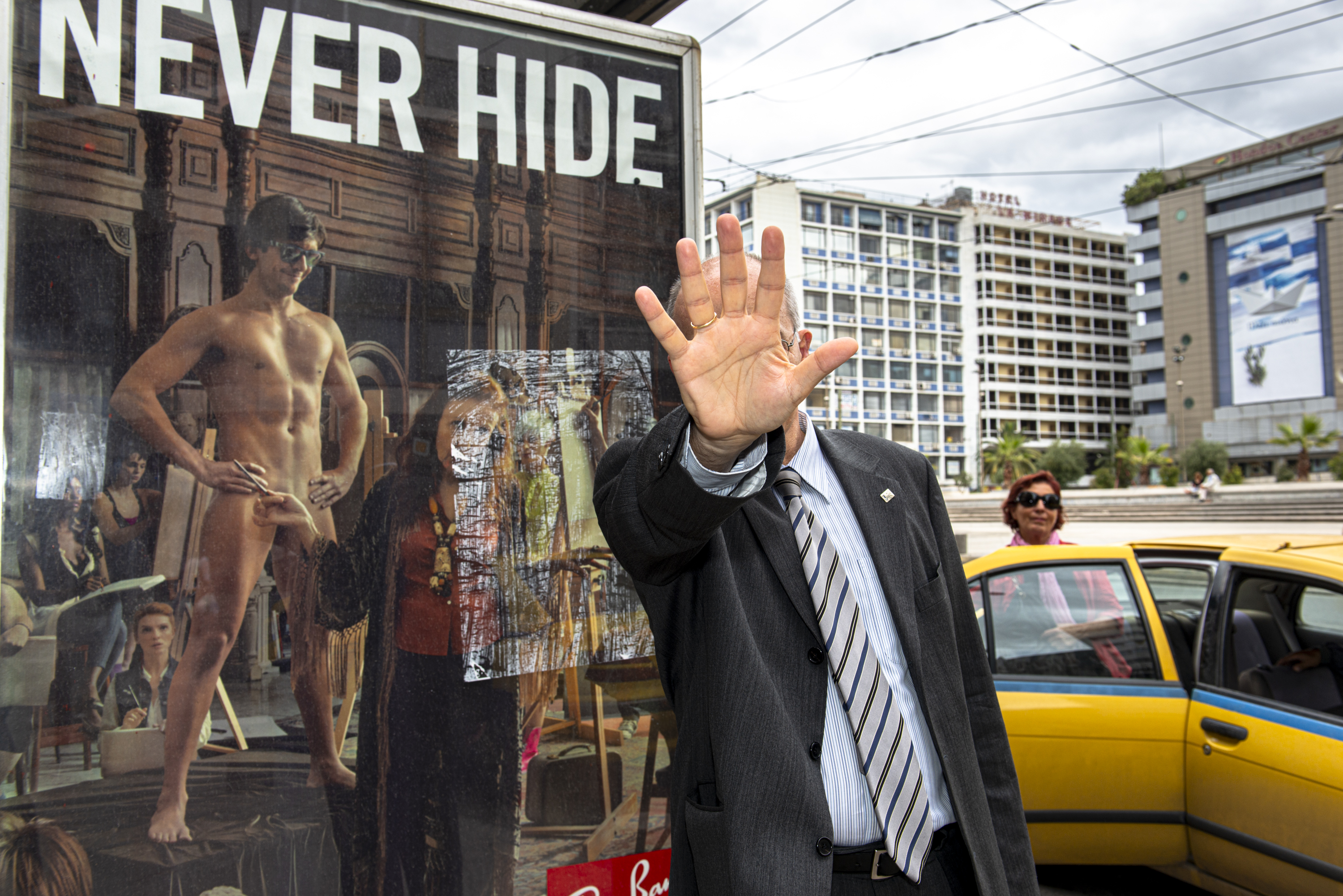 A man in a suit holds his hand up to block a camera in front of a large "Never Hide" advertisement featuring a nude model.
