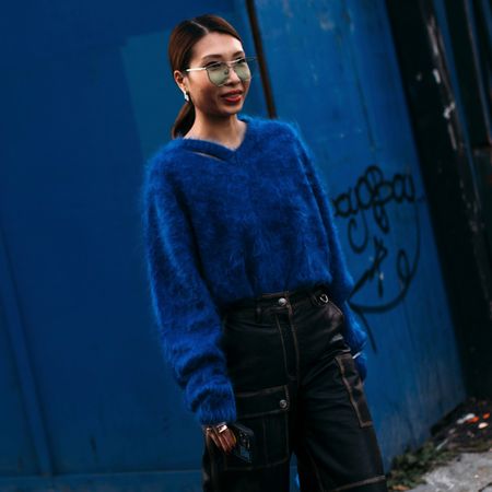 Street style attendee wearing cobalt blue fuzzy sweater black cargo pants, clear sunglasses angainst blue wall 