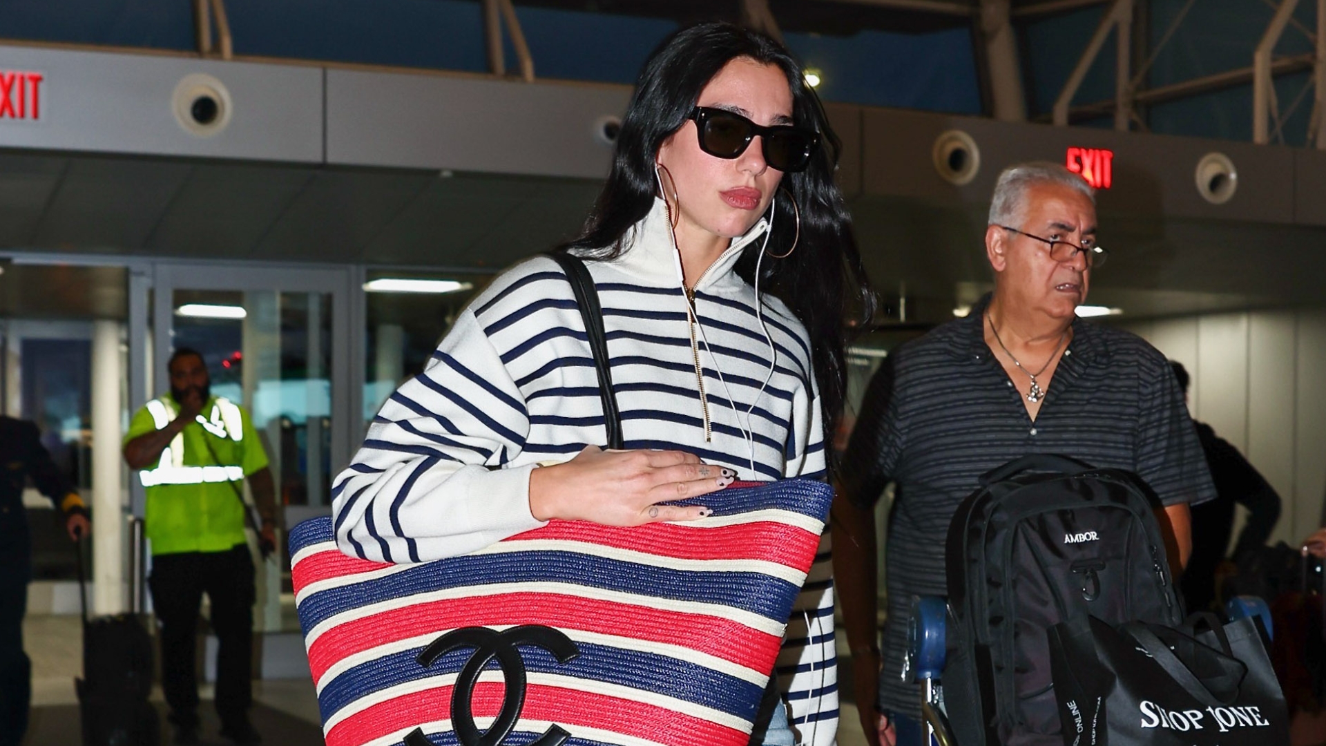 Dua Lipa walks through the airport carrying an oversized Chanel tote bag wearing a striped knit with jeans and black pointed-toe heels.