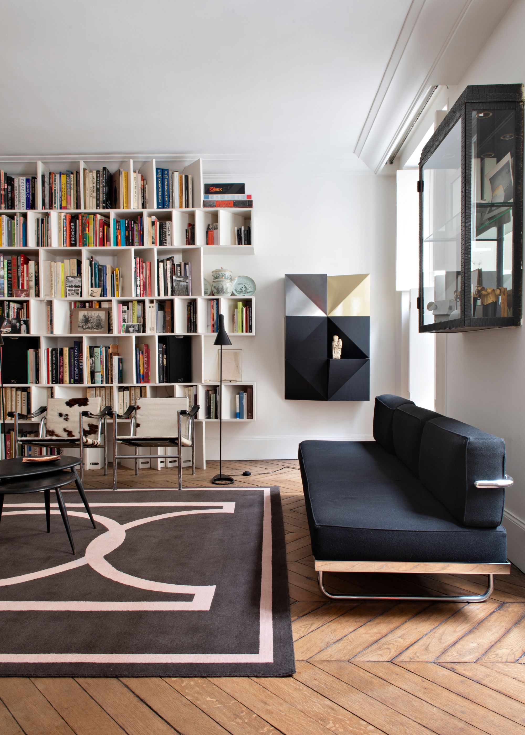 A large living room with white walls, wooden parquet flooring, and a large black and cream rug. A modern and minimal black sofa against the wall and a floor-to-ceiling open shelving unit on the far wall.