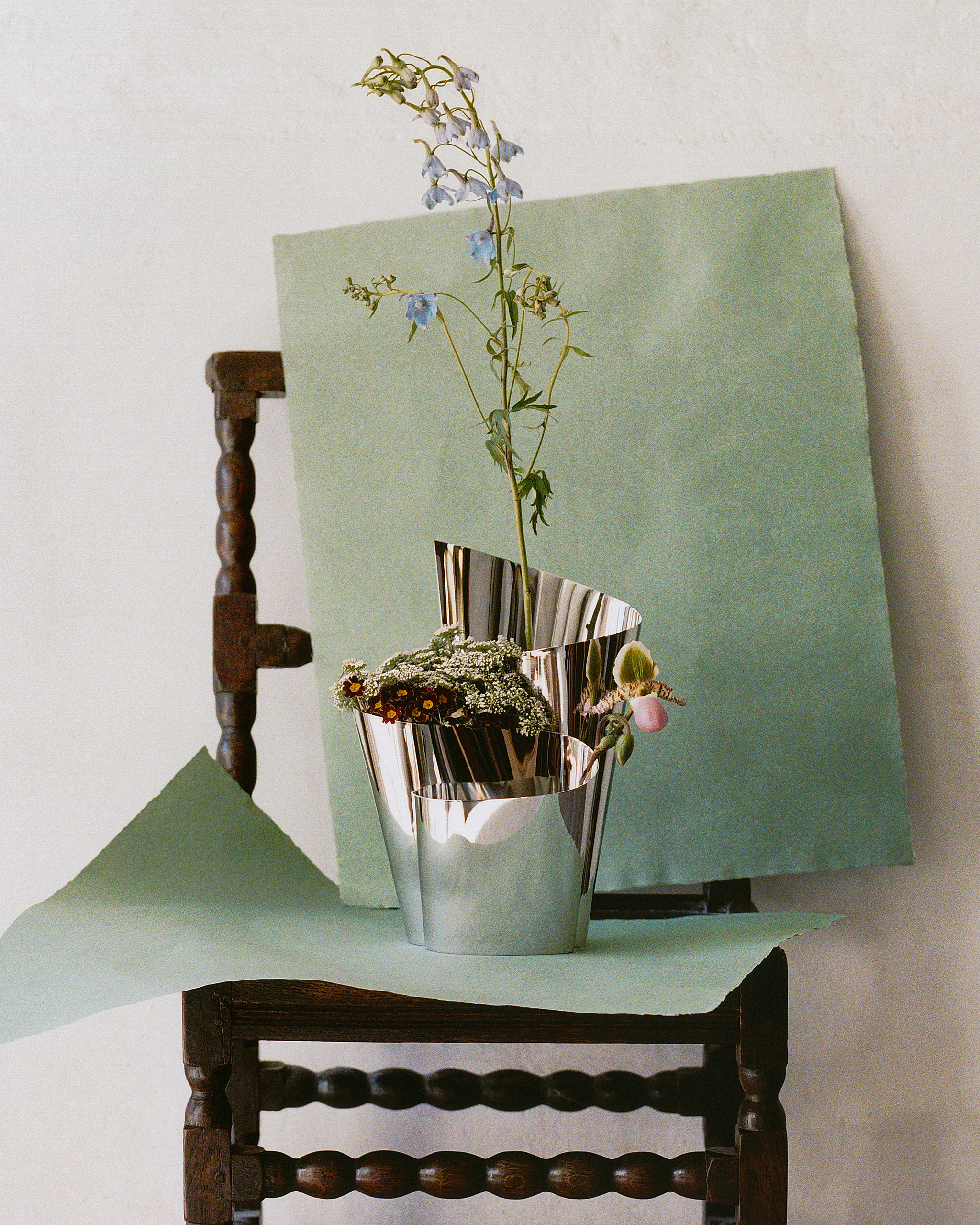 An abstract chrome flower vase with stems in it sitting on a wooden, bobbin chair that has two sheets of sage paper across the seat and back.