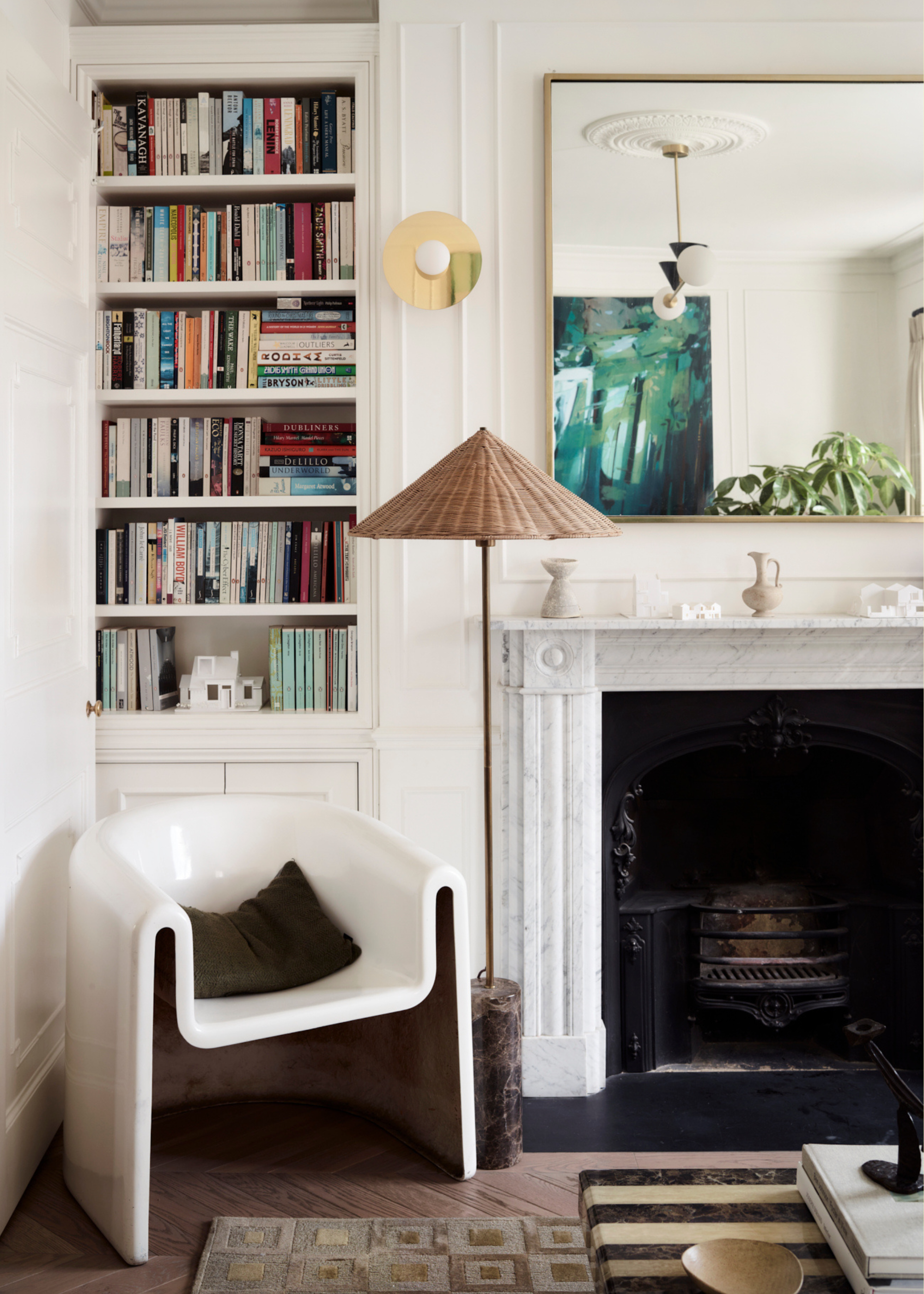 A white living room with a shelf of books, a white armchair, a black pillow, a rattan style lamp with a marble base, a gold wall sconce, a large mirror over a marble mantel, and a black fireplace