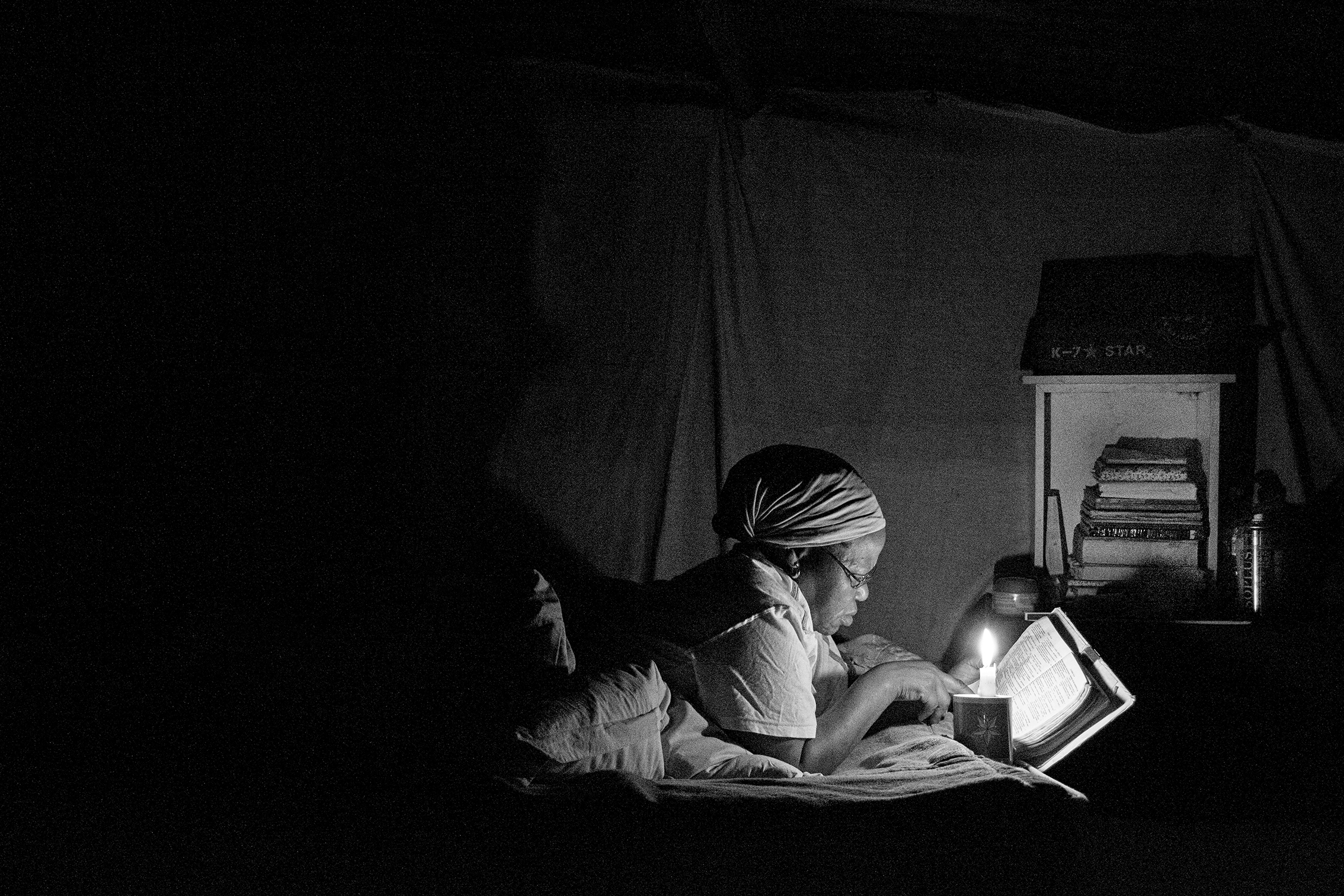 Black and white photograph of a woman lying on her stomach reading a bible by candlelight