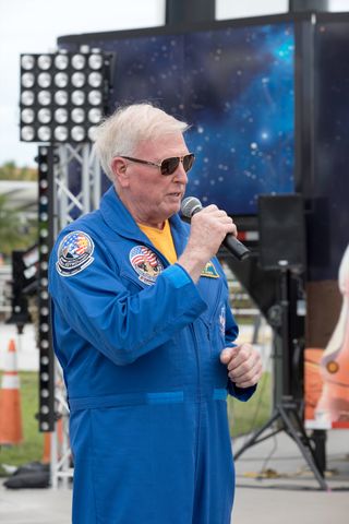 an old man in a blue flight suit and sunglasses speaks into a microphone at an outdoor event