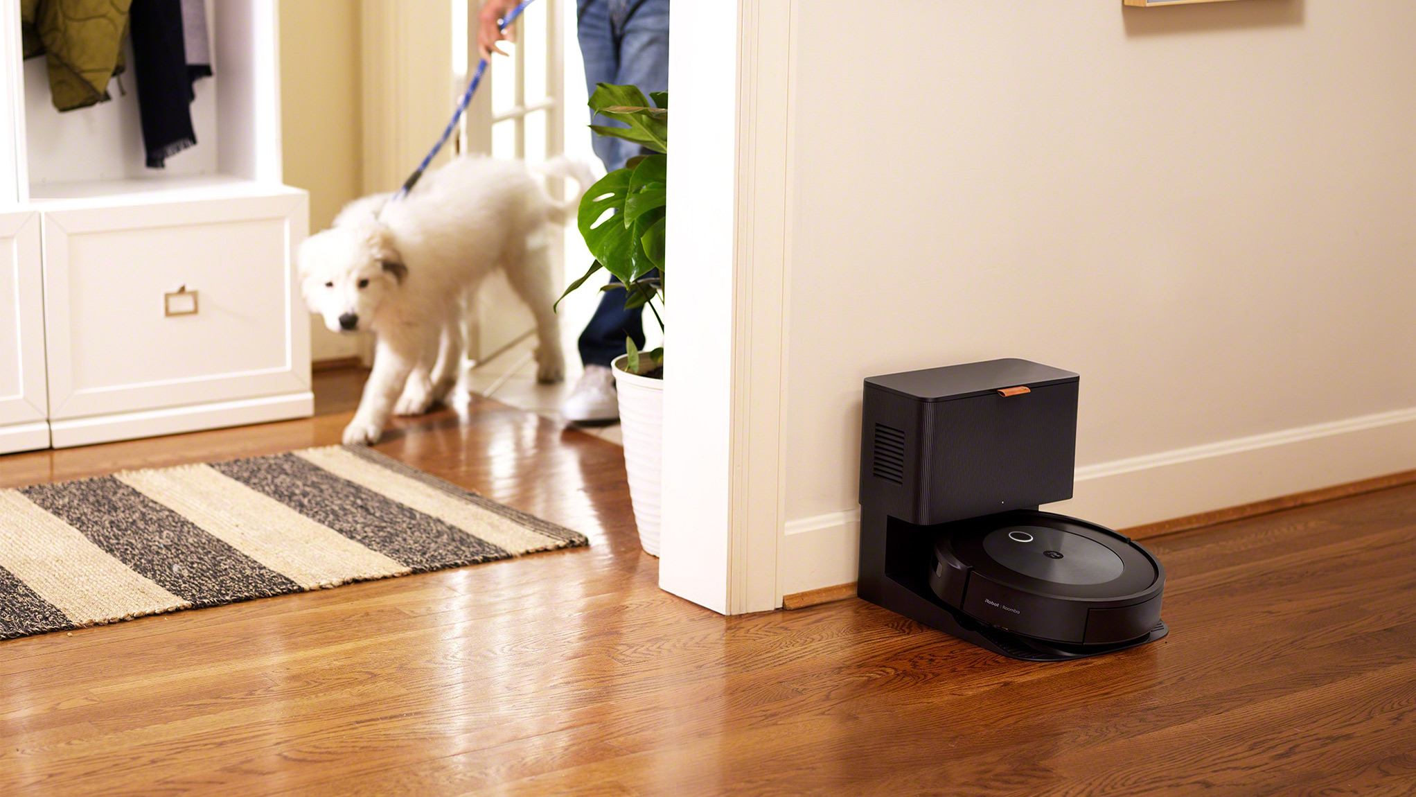 The iRobot Roomba J7+ on its charging station in a hallway while a man with a dog enters the room