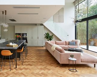 A kitchen extension with a pink corner sofa facing glass sliding doors in front of a dark island with orange bar stools