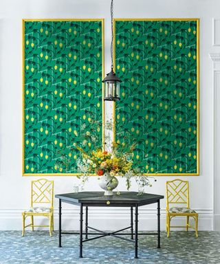 Two large panels with yellow frames, green and yellow botanical wallpaper, white walls, two yellow chairs, black metal side table, terrazzo tiled floor