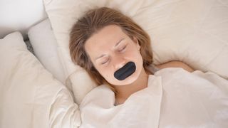 A woman lies asleep on her back in bed, her mouth covered with black mouth tape which she hopes will help her sleep better