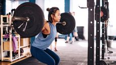 An East Asian woman in a tank top works out in a gym. On her shoulders is a heavy barbell; she's squatting as she pushes herself back up to a standing position.