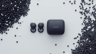 Sony LinkBuds i svart ligger bredvid sitt tillhörande laddningsforal på ett vitt bord, omgivna av svarta små pärlor.