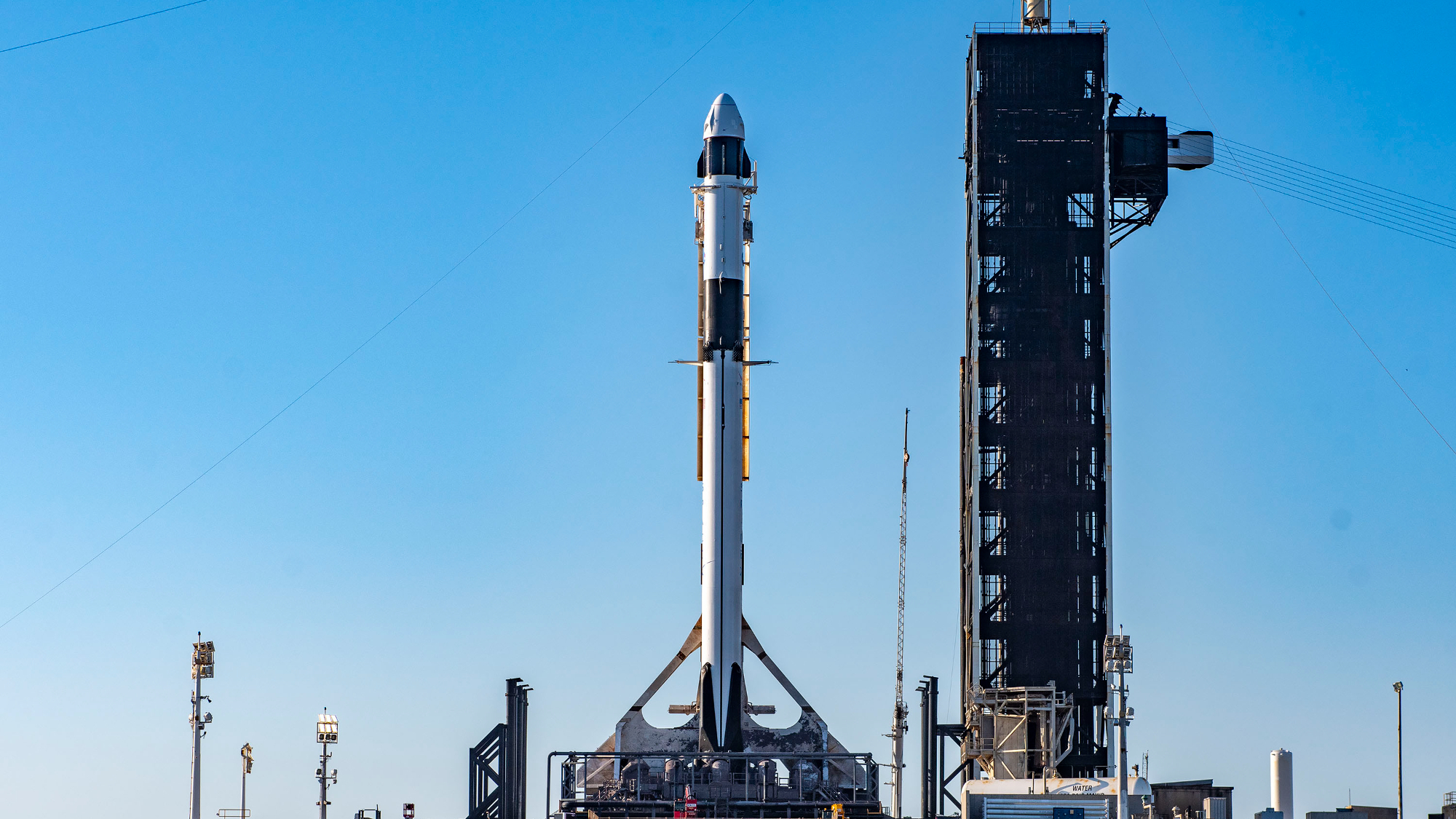 Fusée noire et blanche surmontée d'un support de capsule blanc sur la rampe de lancement avec un ciel bleu en arrière-plan.