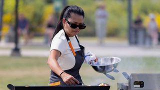 Rhoda cooking on an outdoor grill with an apron and sunglasses on during Top Chef. Background is blurred.