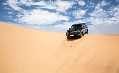 Toyota Land Cruiser coming over a sand dune with a blue sky and clouds behind it.