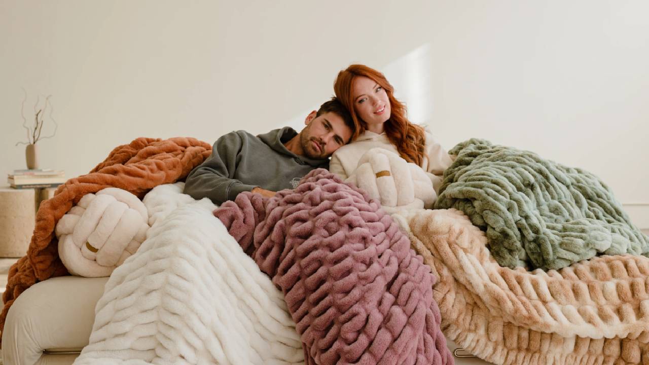 A man and a woman sitting together on a cream sofa, cosied up in various color ways of Lola Blankets.