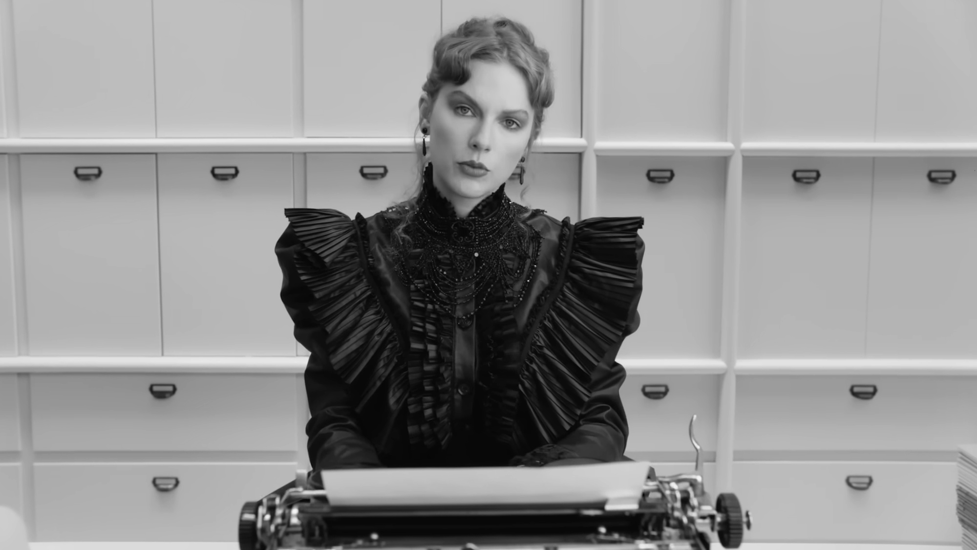 Taylor Swift is shown looking at the camera while wearing a black dress, sitting front of a typewriter in the black-and-white music video for "Fortnight."