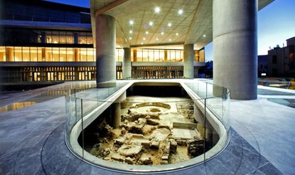 An exterior section of the museum, featuring grey concrete pillars and floors with a capsule shaped hole in the middle containing archeological-like items. The barrier is around the capsule-like hole is made of clear glass