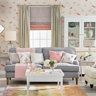 living room with pink and orange dragonfly wallpaper, a window with cream curtains and two tone blinds, a grey sofa with pink and white cushions and a pink throw, and a white coffee table on an orange patterned rug