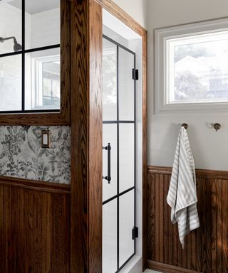 bathroom with dark gray and white floral wallpaper, wooden paneling, black metal framed shower door and window