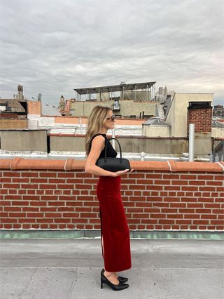 Eliza Huber wearing a black tank top, red velvet midi skirt, black heels, and a black Freja Mercer bag on a roof.