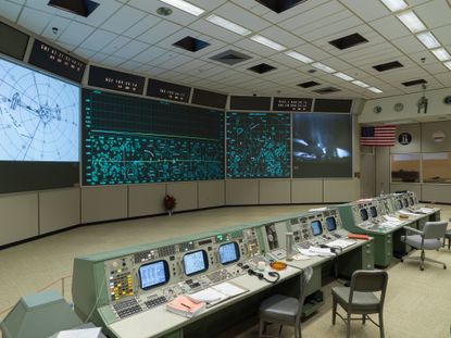 Interior view of the recreated Johnson Space Centre in Houston featuring light coloured flooring, large screens, green workstations with built-in screens, chairs and the American flag