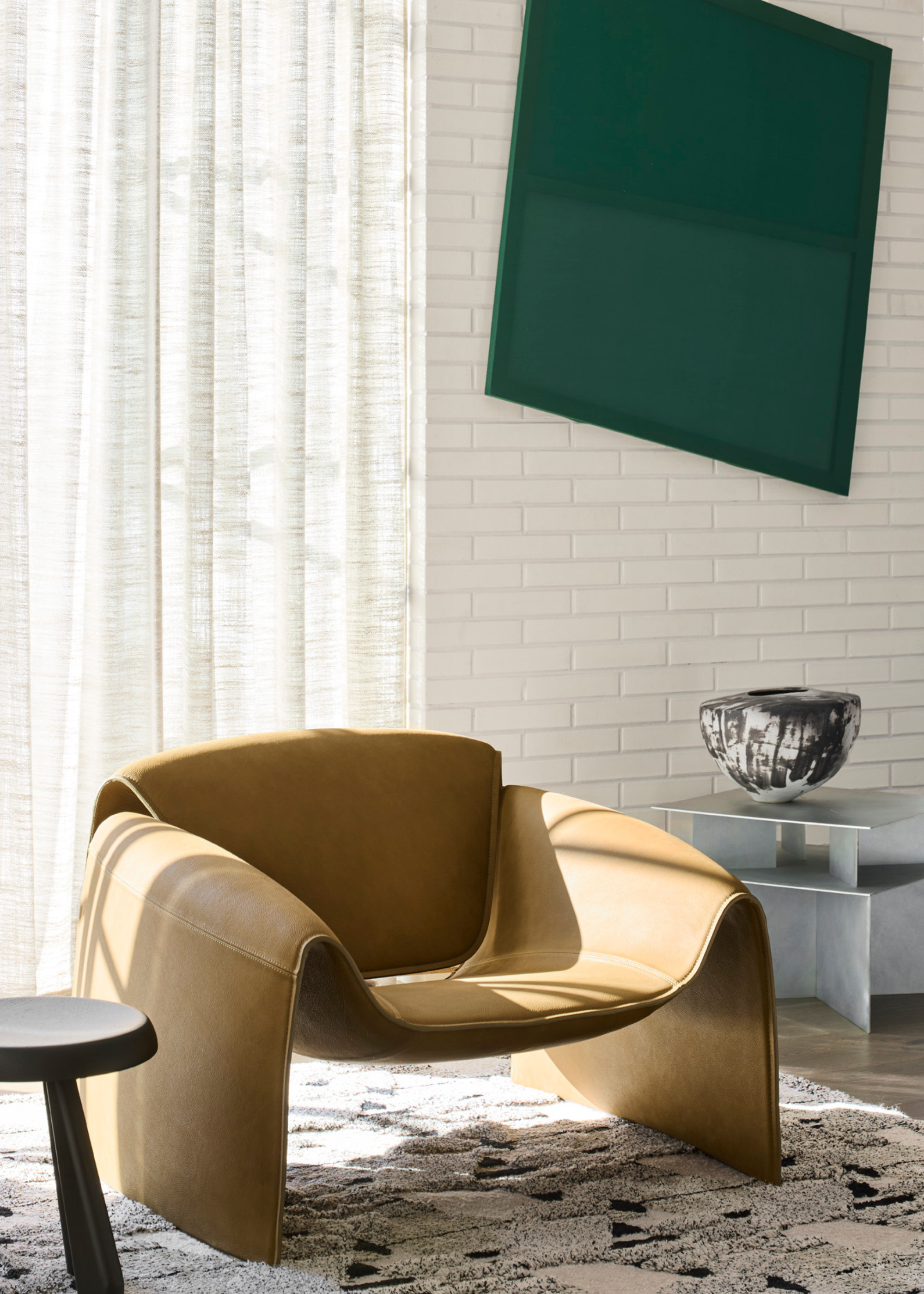 A living toom with a white brick wall, a leaning green art piece, a leather armchair, a wood stool, and a brushed steel side table with a vase on a tufted rug
