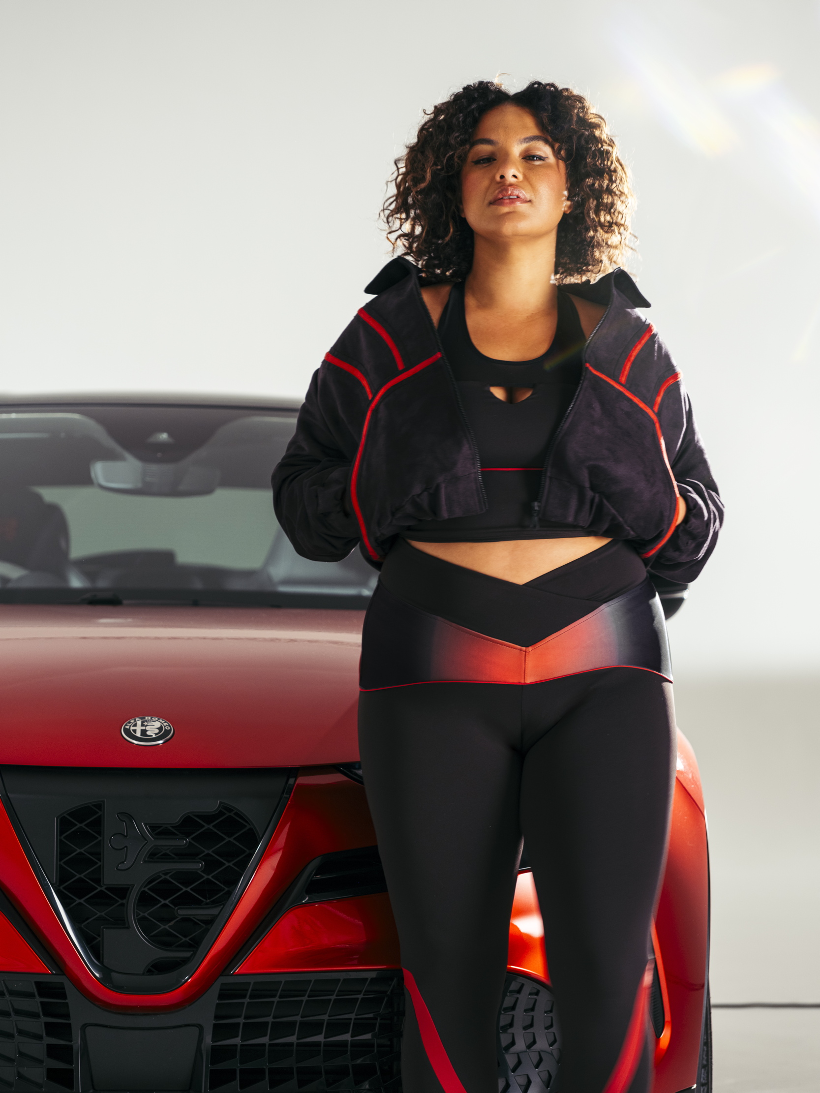 A woman in athleisurewear standing in front of an alfa romeo. the clothes are a stylish mix of red and black