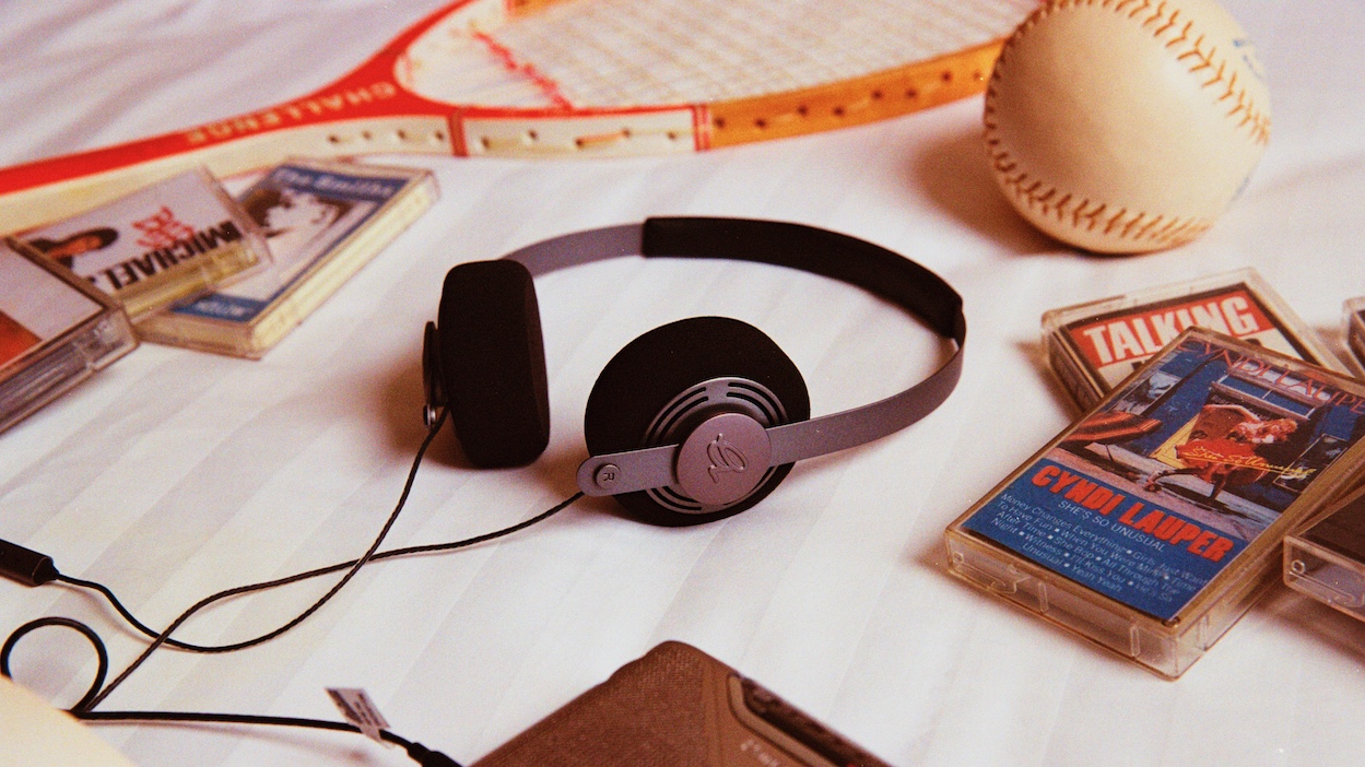 A pair of Gadhouse Wesley retro headphones on a striped surface surrounded by cassette tapes, a tennis racket and baseball.