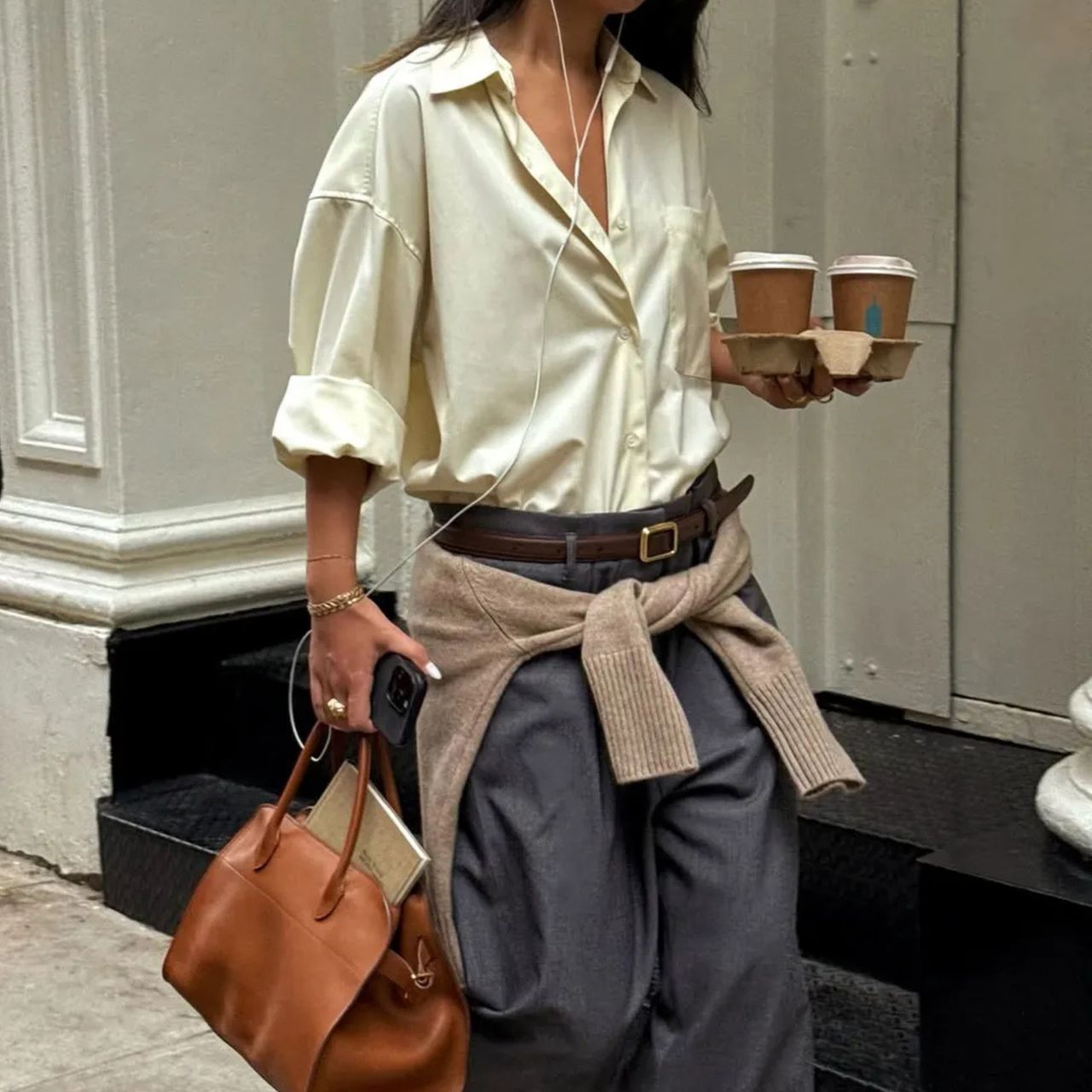 Woman wears grey trousers, yellow shirt and carries a brown The Row Margaux leather tote bag