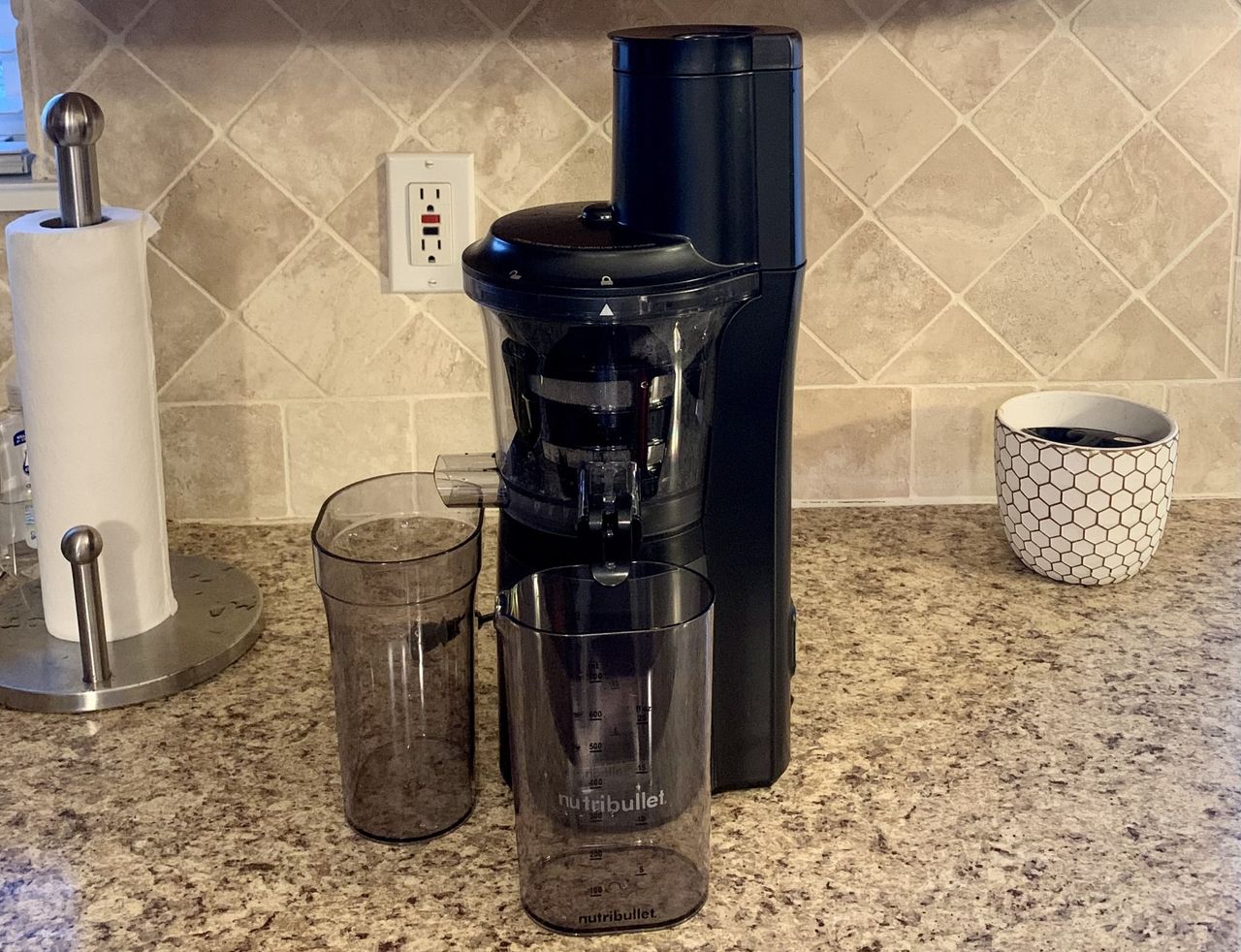 A black cold press juicer with a tall tower compartment on the right hand side, and two transparent grey beakers, one to the left and one in front, on a grey granite kitchen counter.